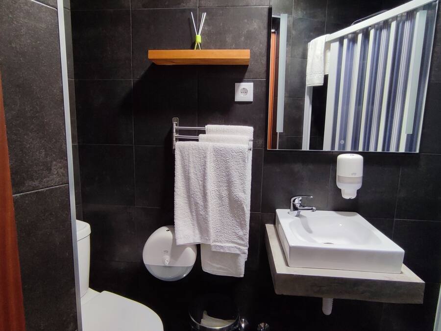 Un baño moderno con inodoro, lavamanos y espejo. Hay un perchero con una toalla, un dispensador de jabón y un pequeño estante con una botella.