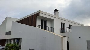 A modern two-story building with a flat roof, a wooden sloped section, and a balcony with a metal railing.