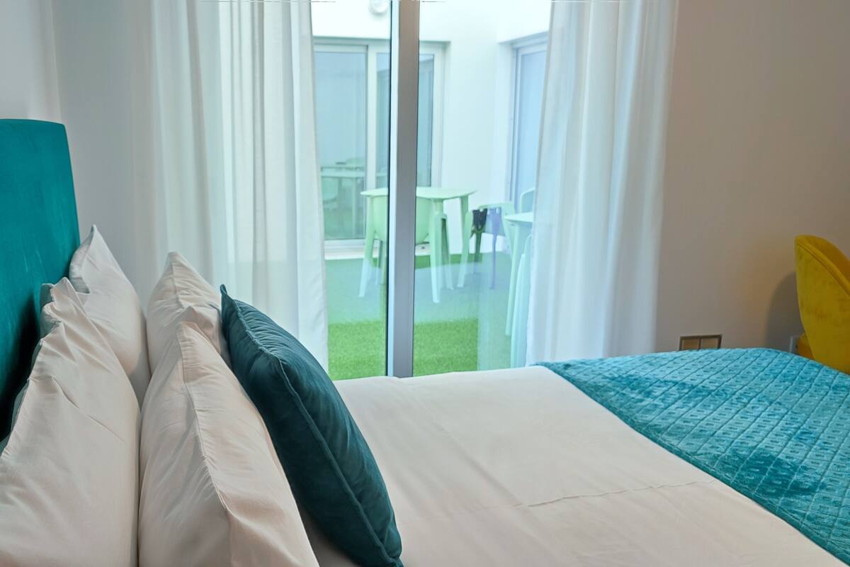 Habitación de hotel con cama, ventana con cortinas y vistas a un espacio exterior con mesas y sillas.