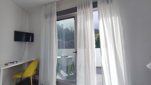 A room with a TV mounted on the wall, a yellow chair, a small table, and a sliding glass door leading to an outdoor area with a table and chairs.