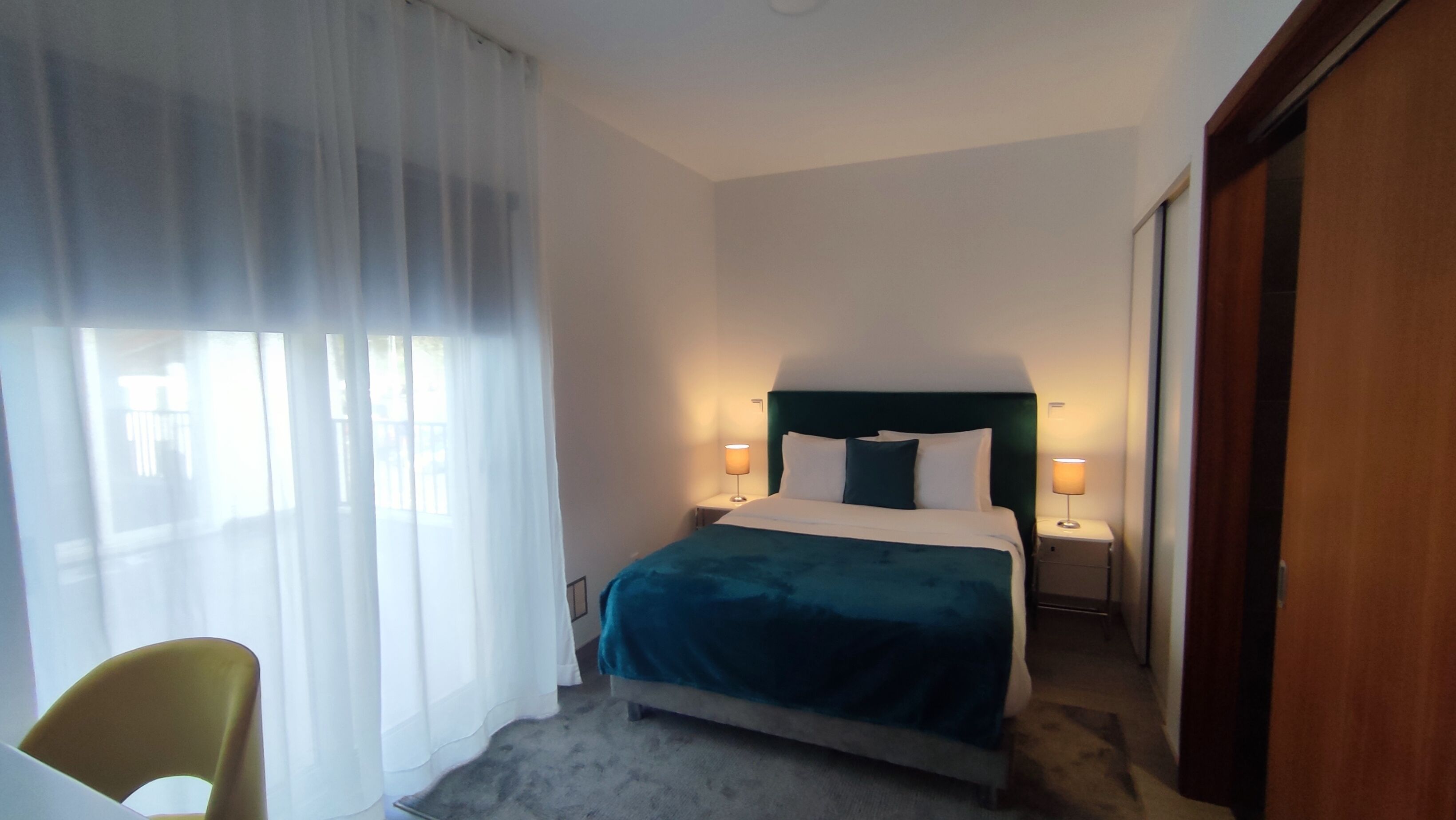 Et hotelværelse med en seng, to sengeborde, en stol og et stort vindue med let gardin.