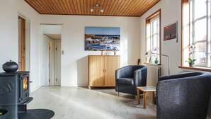 Een woonkamer met een houten plafond, een zwarte kachel, een houten kast, twee blauwe fauteuils, een kleine tafel en een schilderij van een zeegezicht.