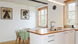 Een keuken met witte kastjes, een houten aanrecht, een spoelbak en een raam met uitzicht op bomen. Er staan twee stoelen bij een tafel met een groene tafelkleed.