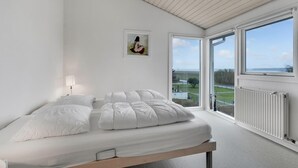Un dormitorio con cama, mesita de noche, lámpara, cuadro enmarco y vistas al exterior a través de amplias ventanas.