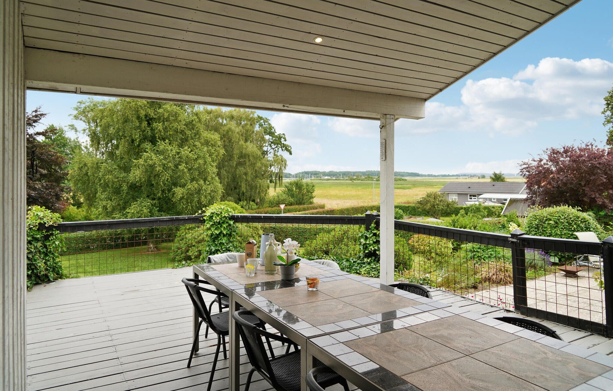Een overdekt terras met een tafel en stoelen, met uitzicht op een groen landschap en een huis in de verte.