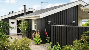 Una casa negra con puerta blanca, chimenea y un pequeño jardín con flores y plantas.