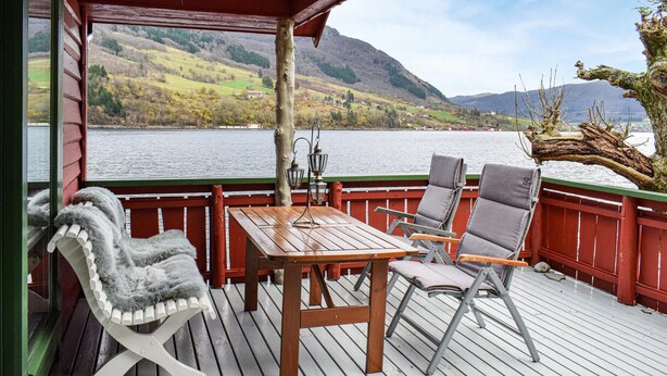 Una terraza de madera con una mesa y dos sillas, con vista a un lago y colinas al fondo.