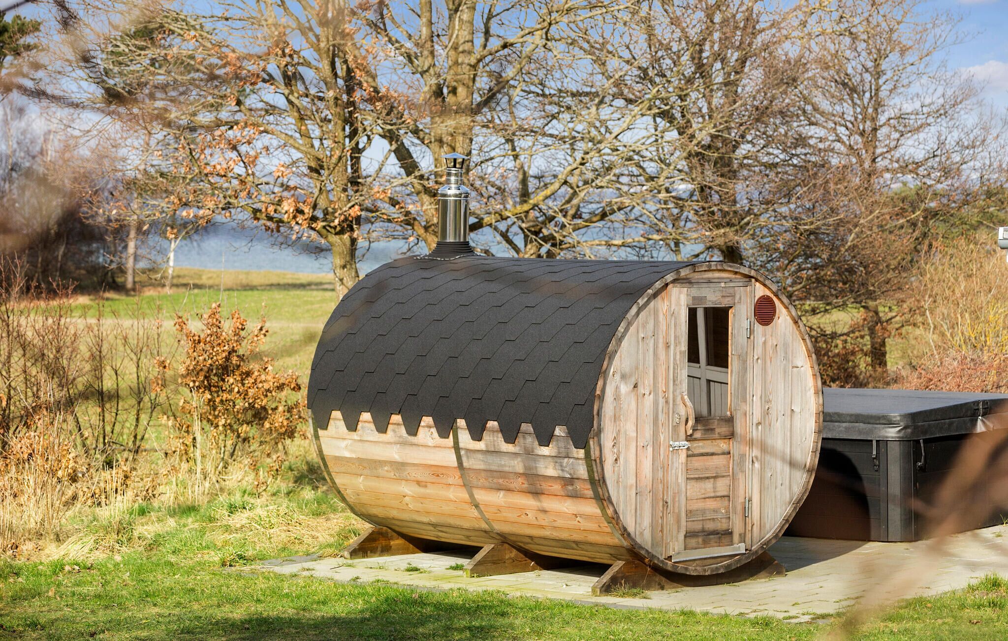 En træ-brændt sauna i tøndestil med dør og skorsten, beliggende udendørs med træer og græs i baggrunden.