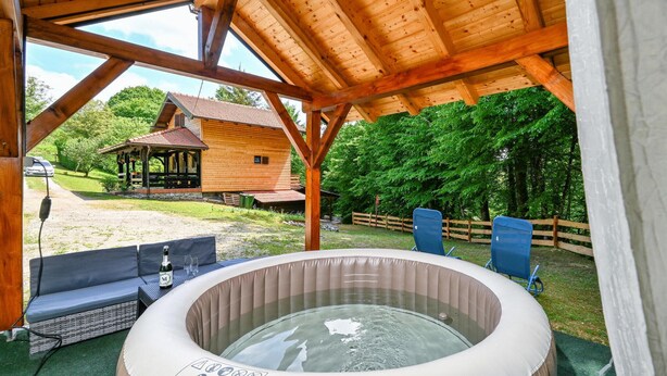 A hot tub under a wooden pergola with a view of a rustic cabin and a fenced garden.