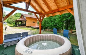 Un jacuzzi bajo una pérgola de madera con vistas a una cabaña rústica y un jardín cercado.