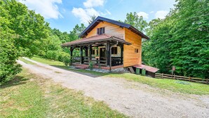 A wooden cabin with a porch, surrounded by lush greenery and a gravel path.