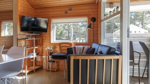 Acogedor interior de cabaña con detalles de madera, un televisor montado en la pared, una zona de comedor con mesa y sillas, y una sala de estar con sofá y ventana con vistas a los árboles.