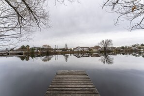 Une jetée en bois s’étendant sur une étendue d’eau calme, où se reflètent des maisons et des arbres sans feuilles.