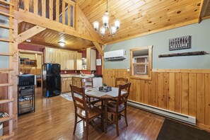 L’intérieur d’un chalet en bois comprend une table à manger, des chaises, un lustre, un réfrigérateur et un coin cuisine.
