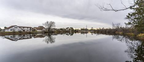 A calm lake with a reflection of buildings and trees, surrounded by a cloudy sky.