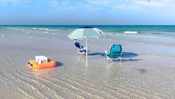 A beach with clear skies, a floating orange object, two beach chairs, and a striped umbrella. - Exceptional Condo on the Bradenton Riverwalk (Bradenton)