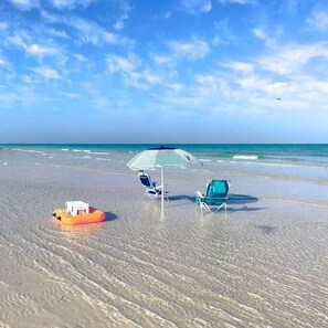 A beach with clear skies, a floating orange object, two beach chairs, and a striped umbrella. - Exceptional Condo on the Bradenton Riverwalk (Bradenton)
