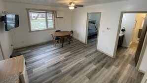 Une pièce dotée d’un plancher en bois, d’un ventilateur de plafond, d’un téléviseur fixé au mur, d’une fenêtre et d’une porte donnant sur une autre pièce.