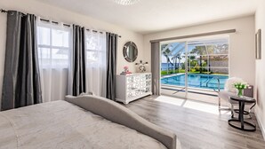 Ein Schlafzimmer mit einem großen Bett, einem Schminktisch und Blick auf ein Schwimmbecken durch eine Glastür.