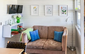 Een woonkamer met een bruine bank, een aan de muur bevestigde televisie en lijstwerken met plantenmotieven.