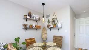 A dining area with a glass table surrounded by wicker chairs, a hanging light fixture, and shelves with decorative items.