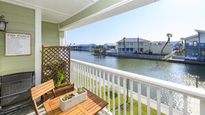 A balcony with a wooden table and chairs, a white railing, and a view of a canal with buildings across it.