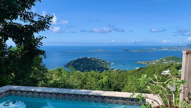 Une piscine dont le bord est revêtu de carrelage, entourée d’une végétation luxuriante et offrant une vue imprenable sur la côte, avec ses îles et son ciel bleu azur.