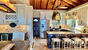 Une cuisine spacieuse avec des meubles blancs, un grand îlot central doté de tabourets, un ventilateur de plafond et un panneau d’accueil.