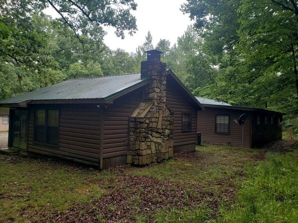 A log cabin with a stone chimney, surrounded by dense forest.