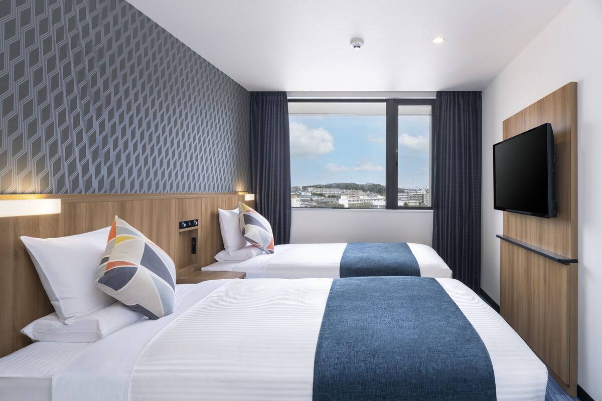 A hotel room with two beds, a TV, a window with a city view, and a patterned wall.
