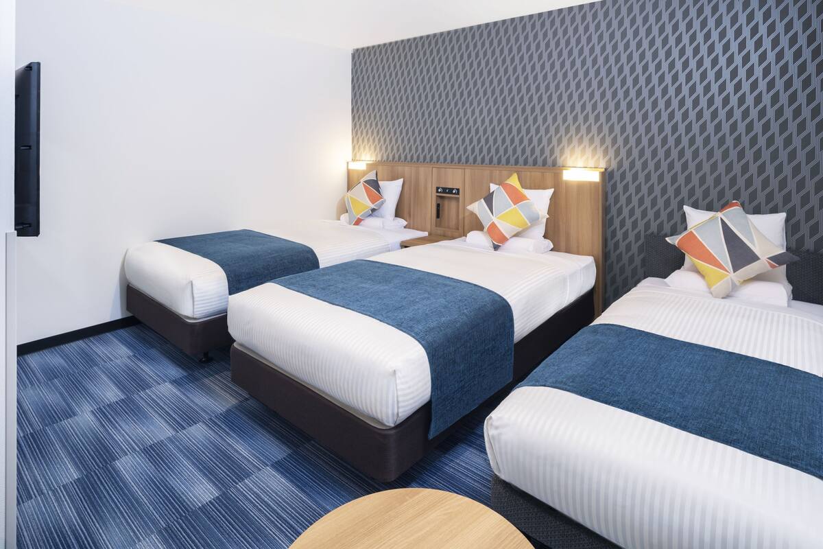A hotel room with three beds, each with a blue bedspread and white linens. There is a wooden headboard with a geometric pattern, and the walls have a similar design. A small table is visible in the foreground.
