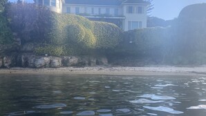 Une propriété située au bord de l’eau, avec une plage, une grande maison dotée d’un balcon donnant sur l’eau et une falaise rocheuse.