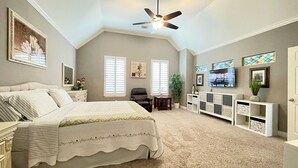 A bedroom with a large bed, a ceiling fan, a TV, and a window with blinds.