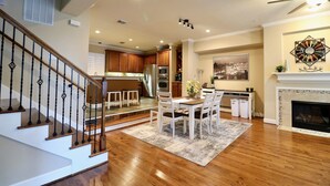 A spacious living area with a wooden staircase, a dining table with chairs, a fireplace, and a kitchen in the background.