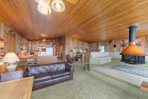 A rustic cabin interior with a wooden ceiling, a leather sofa, a fireplace, and a kitchen area.