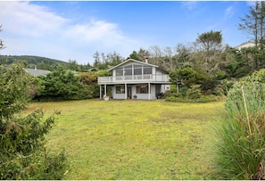 A two-story house with a balcony, surrounded by a grassy yard and dense vegetation.
