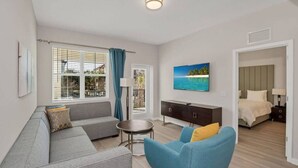 Ein modernes Wohnzimmer mit grauem Sofa, blauem Sessel, kleinem runden Tisch und Fernseher an der Wand. Im angrenzenden Zimmer befindet sich ein Bett mit weißem Kopfteil.