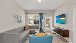 Ein modernes Wohnzimmer mit grauem Sofa, blauem Sessel und einem kleinen runden Tisch. Ein Fernseher steht auf einer Holzkommode und an der Wand hängt ein Bild vom Strand.