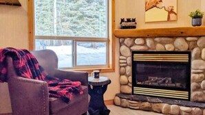 Una sala de estar acogedora con chimenea, un ventanal con vistas a árboles cubiertos de nieve y un sillón cómodo.