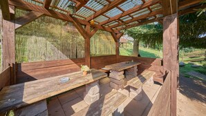A wooden gazebo with a corrugated metal roof, stone benches, and a small table with a flower vase. - Chlívce u Hronova by Interhome (Chlivce)