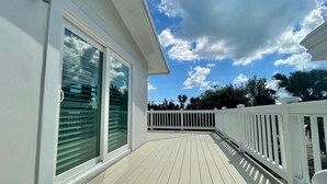A covered outdoor deck with a white railing, a glass door with blinds, and a clear sky with clouds. - Luxury and Lovely Water Front House (Big Pine Key)
