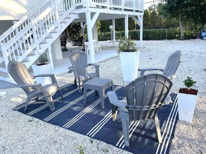 An outdoor seating area with Adirondack chairs, a small table, and a blue striped rug on a gravel surface.