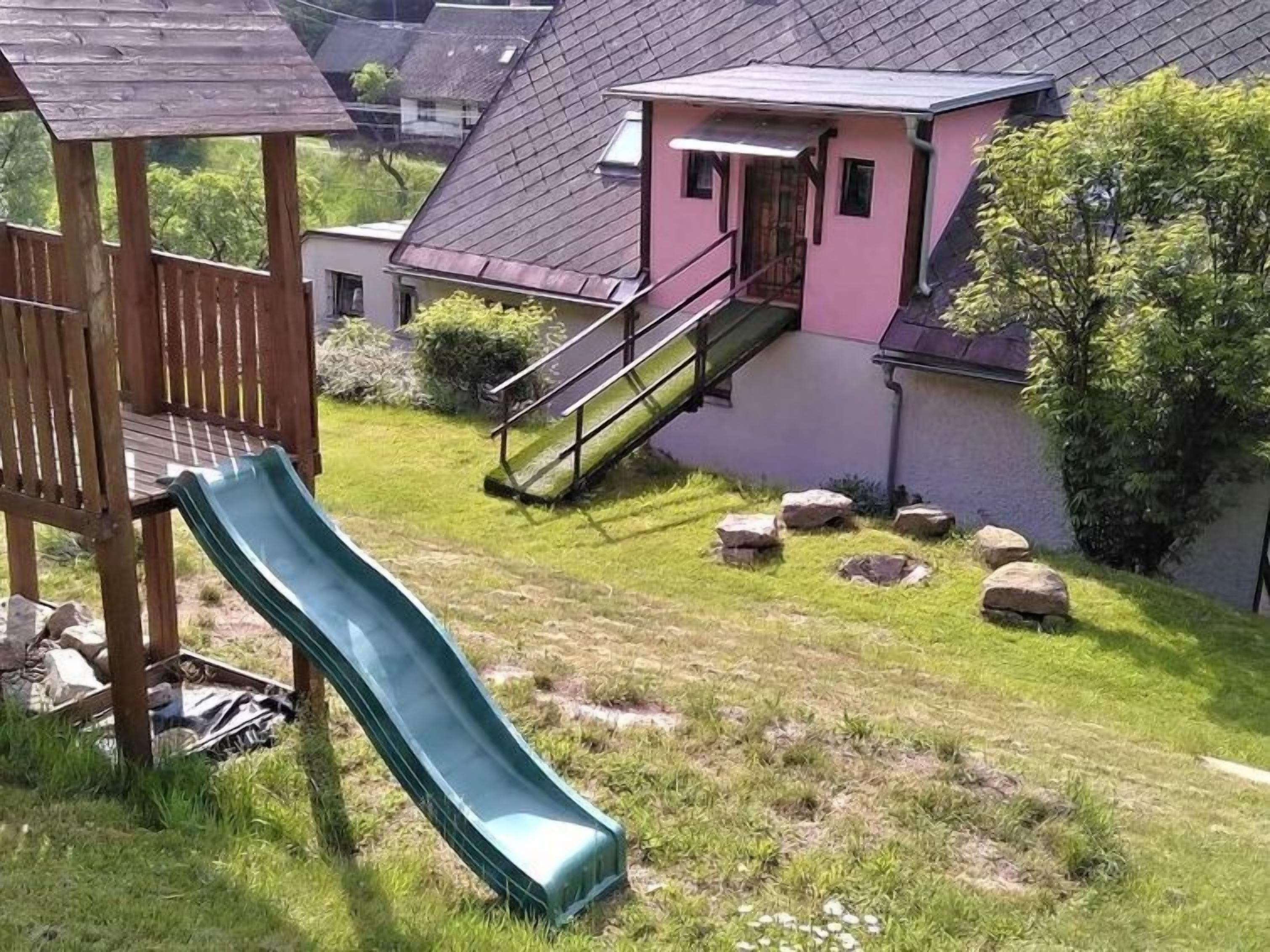 Un pati amb un tobogan verd, una terrassa de fusta i una casa amb façana rosa i teulat gris.