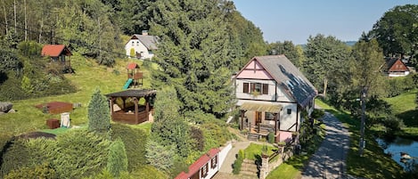 A large house with a sloped roof, a small wooden playhouse, and a pond.