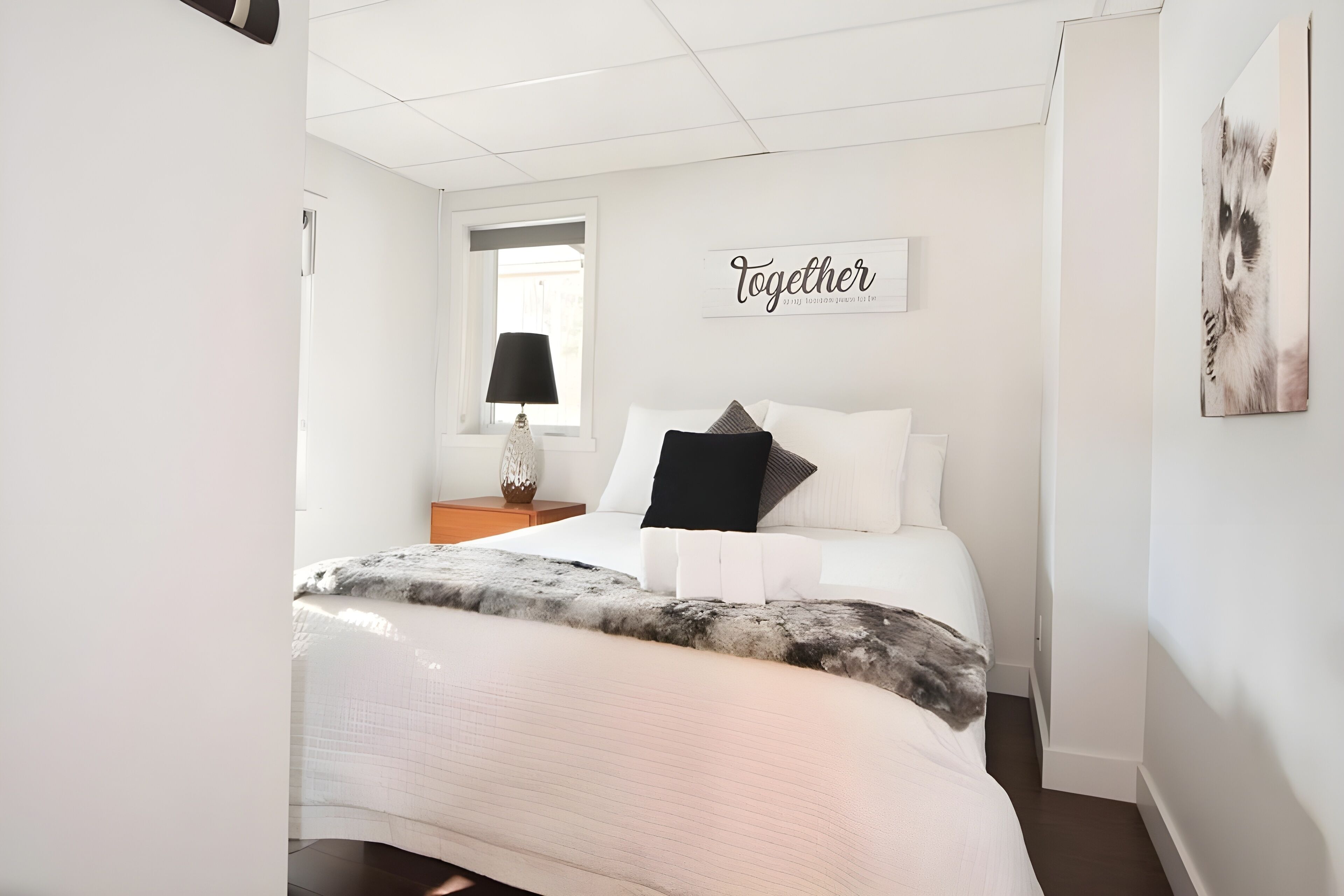 Ein ordentlich eingerichtetes Schlafzimmer mit einem Bett, einem Nachttisch mit Lampe, einem Fenster mit Jalousien und einer Wand mit der Aufschrift 'Together'.