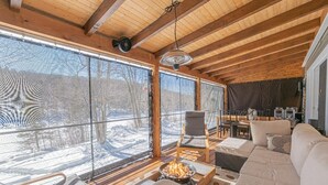 L’intérieur chaleureux d’un chalet, avec une grande fenêtre donnant sur un paysage enneigé, une cheminée et un coin repas.