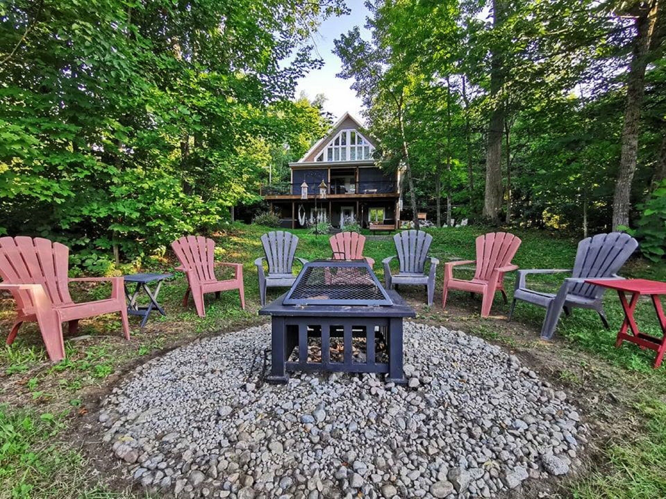 A patio with Adirondack chairs and a fire pit surrounded by gravel, with a house in the background.
