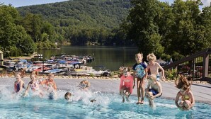 A swimming pool with people enjoying the water, surrounded by a dock with boats and a lake in the background.