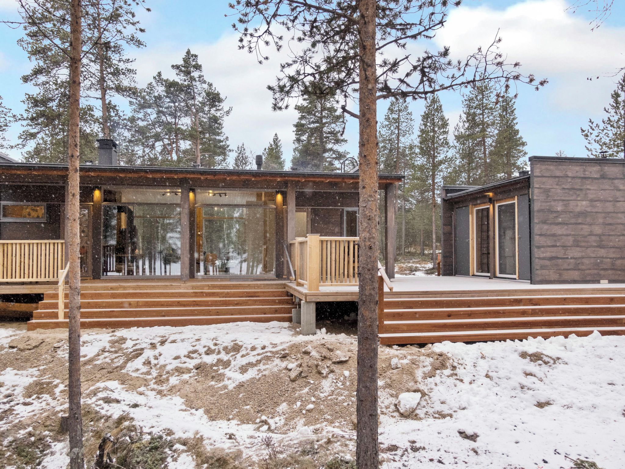 A cabin with a wooden deck and glass doors, surrounded by snow and trees.