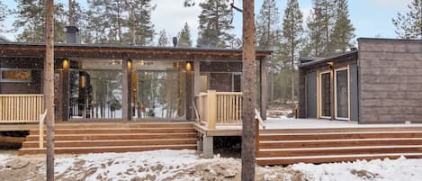 Una cabaña con terraza de madera y puertas de cristal, rodeada de nieve y árboles.